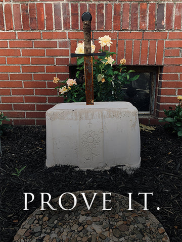 A rusted medieval-style sword is embedded in a white concrete block engraved with a sunflower. The stone sits in mulch with blooming roses in the background. White text at the bottom reads: "PROVE IT."